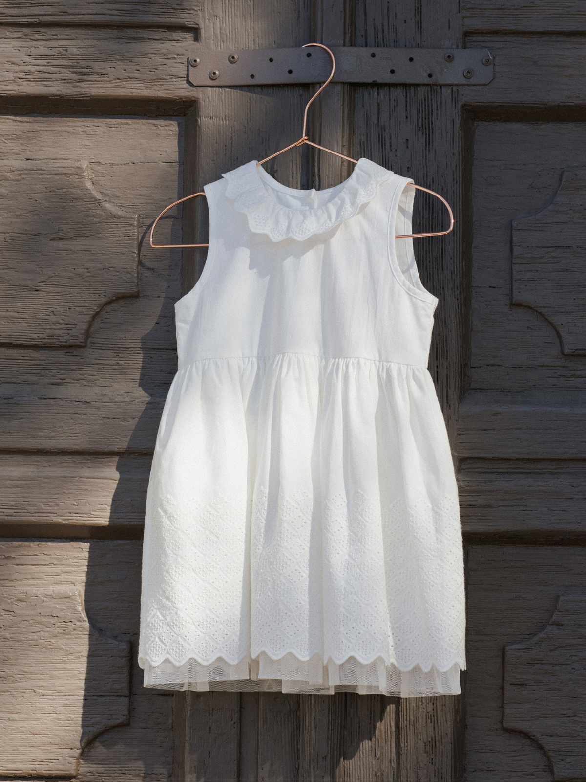 White children's dress with a scalloped collar, hanging on a wooden backdrop.