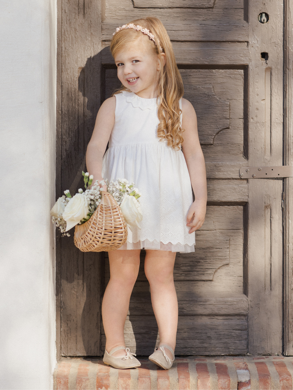 Smiling girl in a white dress holding a woven basket of flowers, standing by a rustic wooden door.
