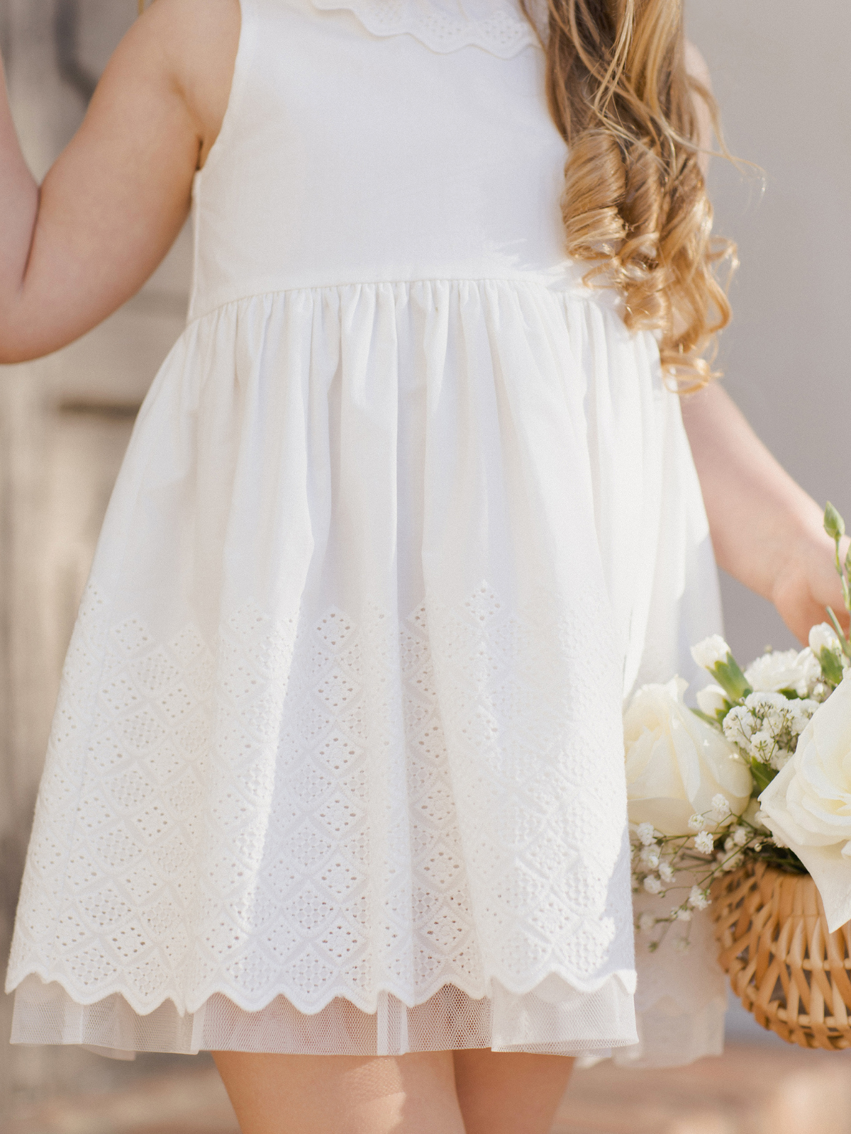 Child in a white dress with lace trim, holding a basket of flowers, standing against a light background.