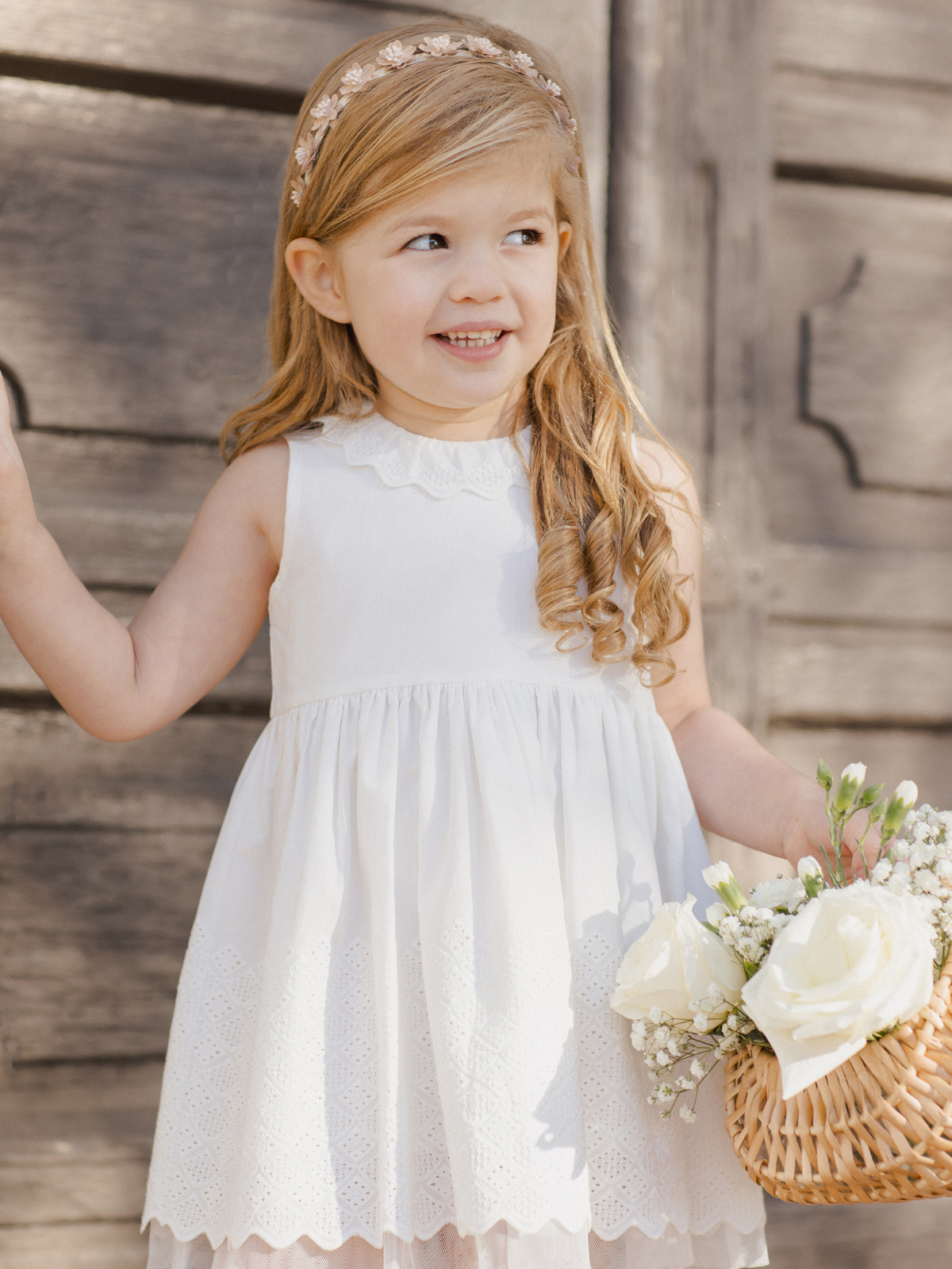 Smiling girl in a white dress holding a basket of flowers near a wooden backdrop.