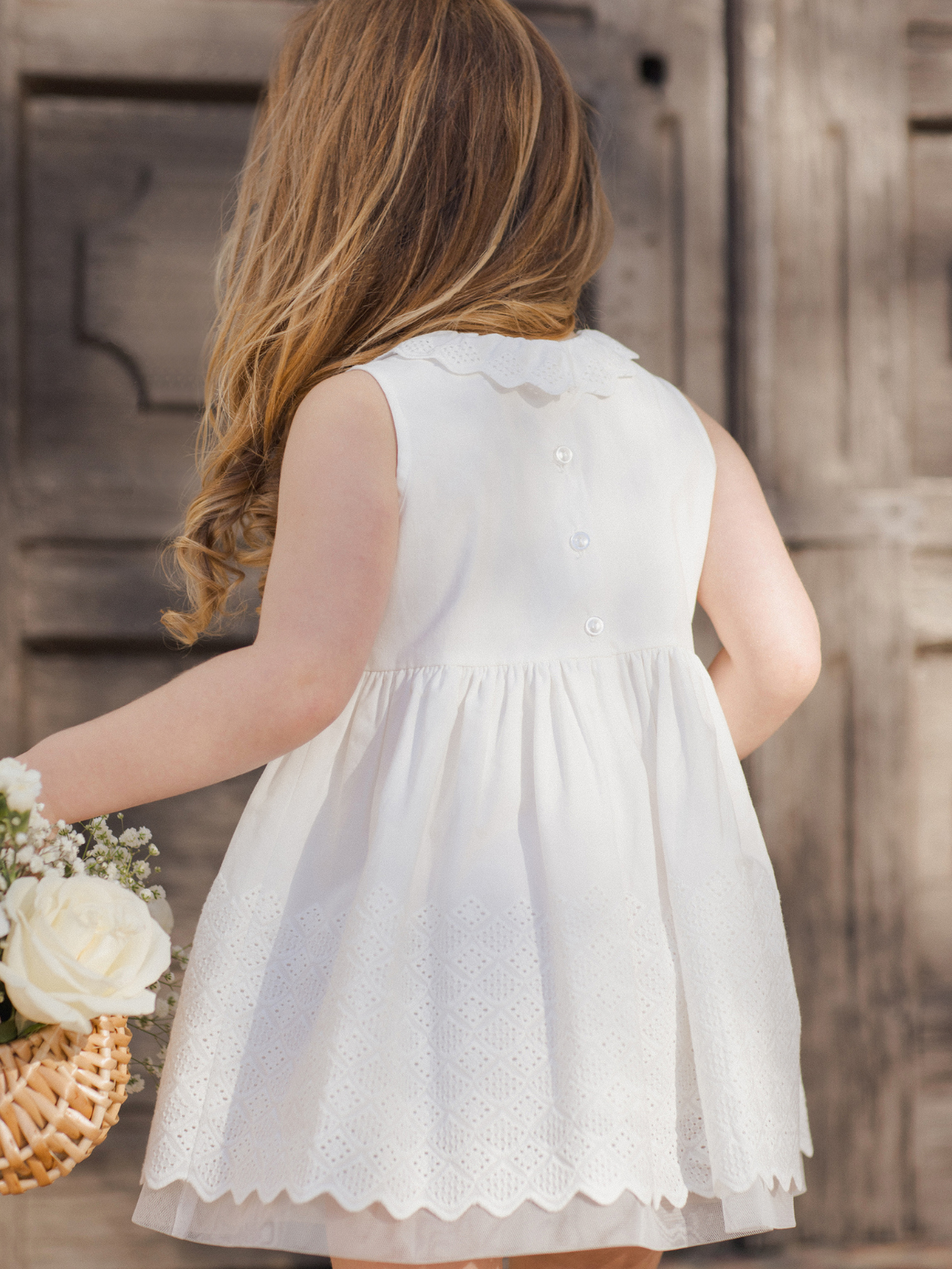 Girl with long hair in a white dress, holding a floral basket, standing in front of wooden doors.