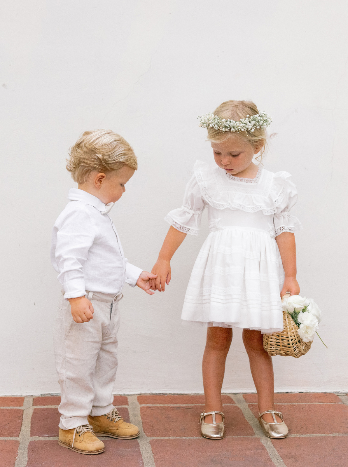 Two children holding hands: a boy in a white shirt and beige pants, and a girl in a white dress with a flower crown.