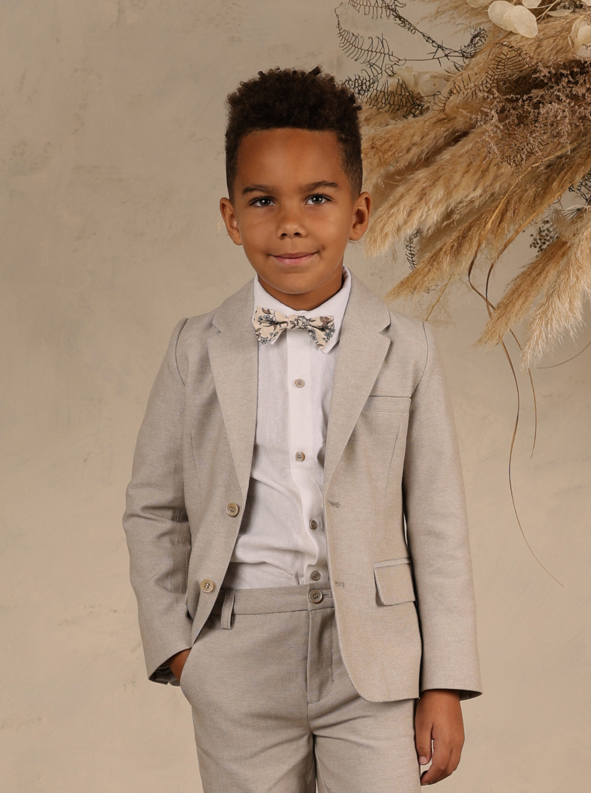 Boy in a light gray suit and floral bow tie, smiling against a neutral background with dried flowers.