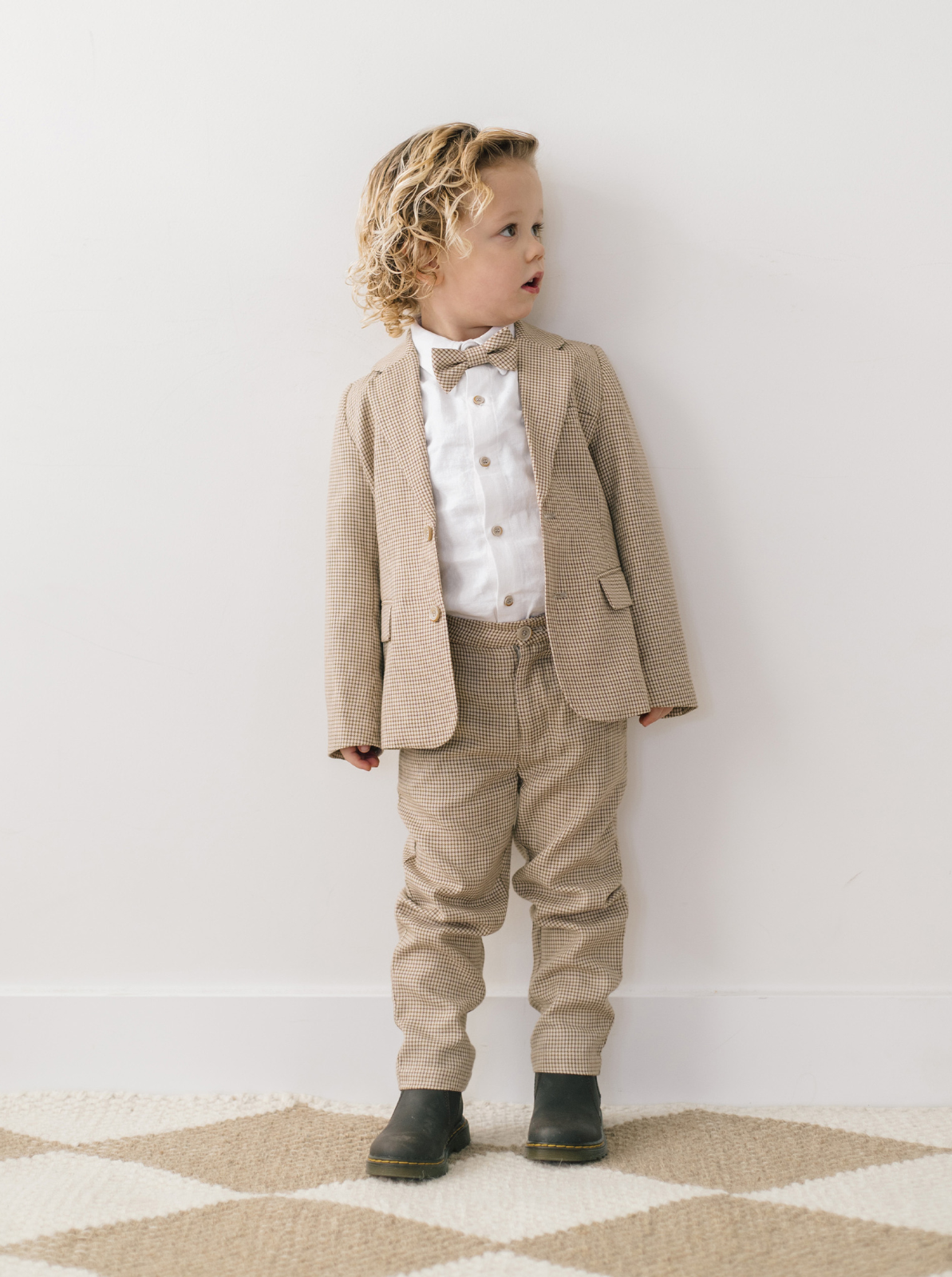 Young boy in a stylish tan suit with a bow tie, standing against a white wall.
