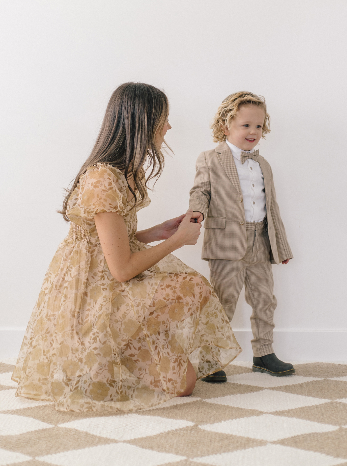 Mother kneels and holds hands with a boy in a suit, both smiling, in a bright, stylish interior.