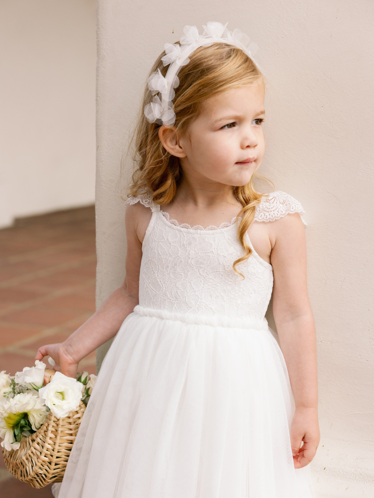 Young girl in a white lace dress and headband, holding a basket of flowers, standing against a light wall.