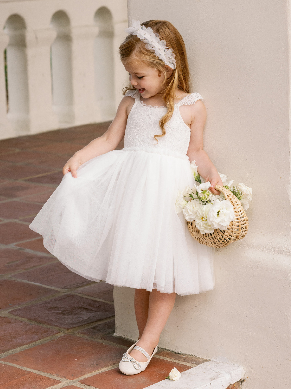 Young girl in a white dress holds a flower basket, smiling, against a light wall in a charming outdoor setting.