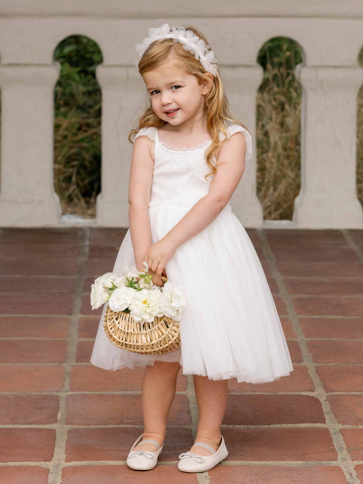 Young girl in a white dress holding a basket of flowers, smiling against a decorative wall backdrop.