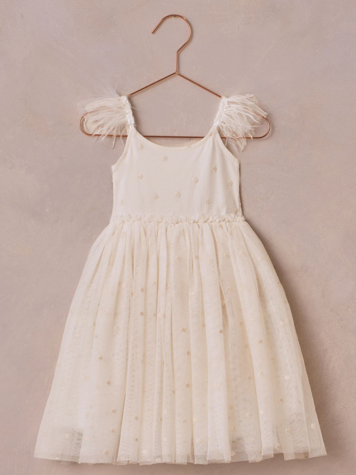Light-colored girls' dress with sheer layers and fluffy straps, hanging on a copper hook against a textured background.