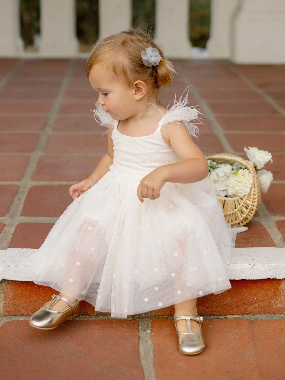 A young girl in a white dress with feathers sits on a patio, wearing gold shoes and a gray hair accessory.