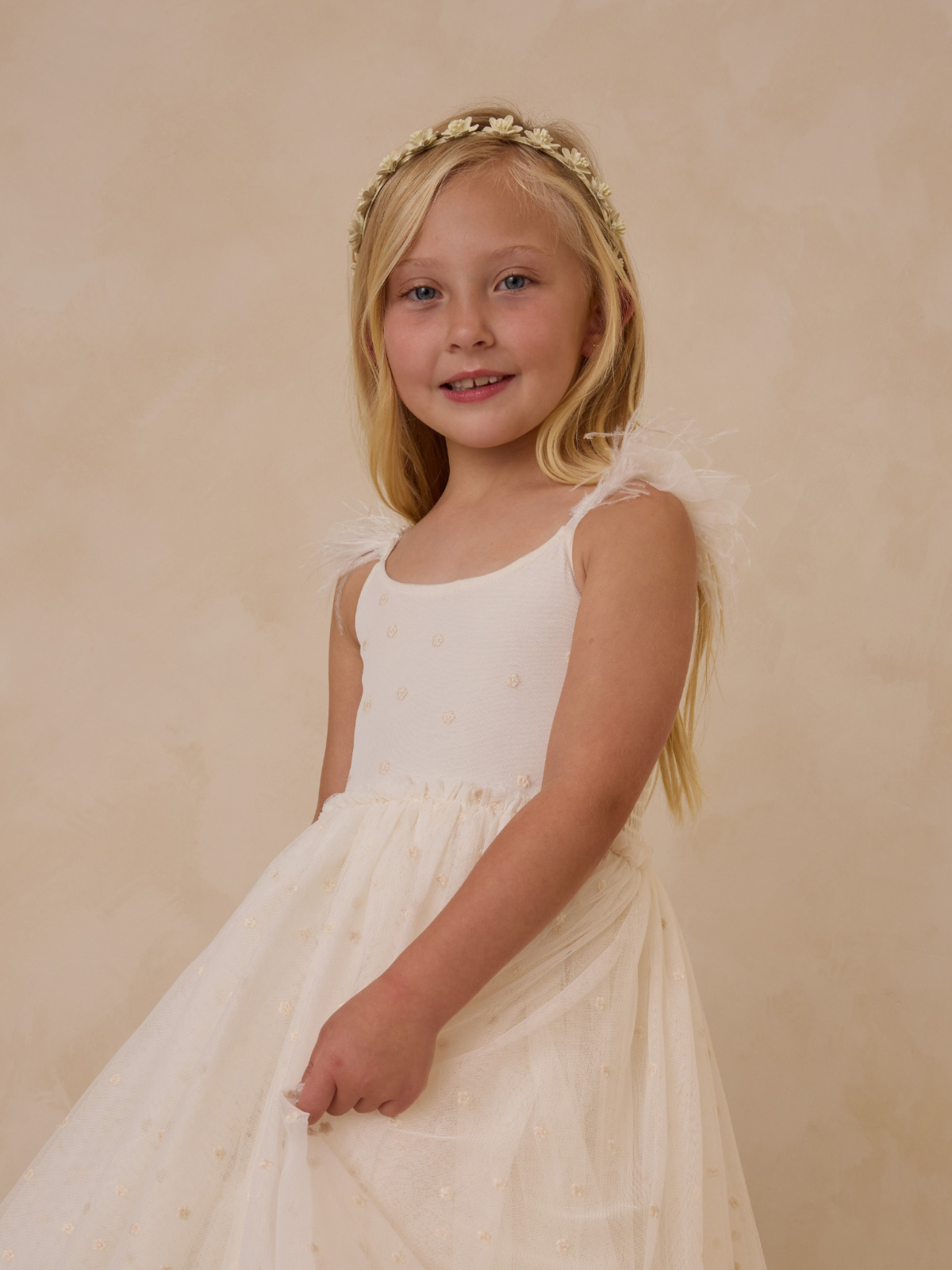 Young girl in a white dress with floral details and a matching headband, smiling against a soft background.