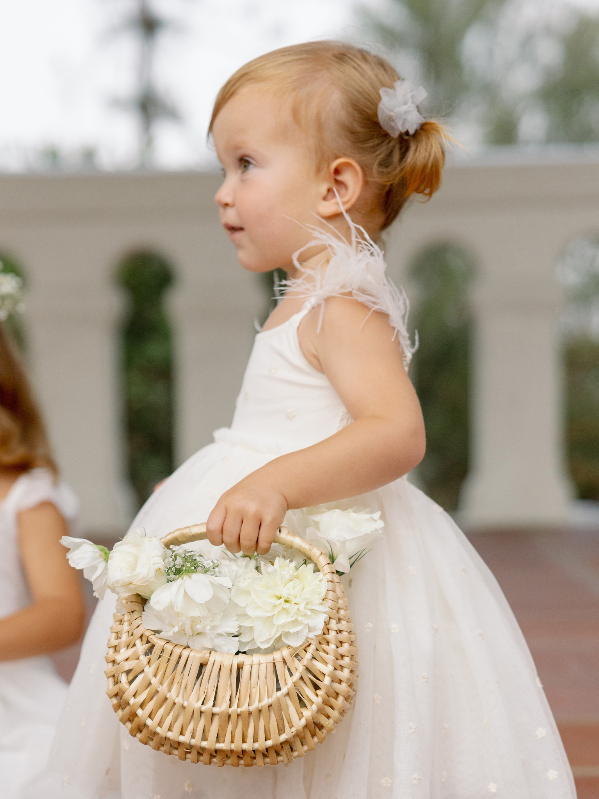A young girl in a white dress holds a basket of flowers, standing in an outdoor setting.