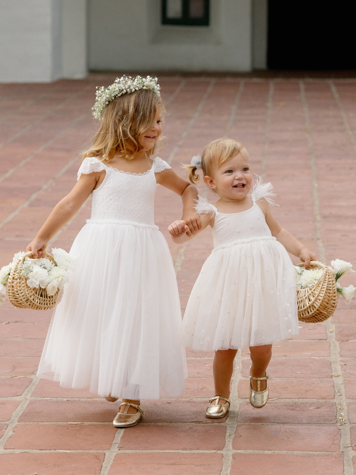 Two young girls in white dresses, joyfully holding hands while carrying flower baskets.