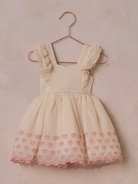 Light cream dress with pink heart embroidery, hanging on a copper-colored hook against a soft pink background.