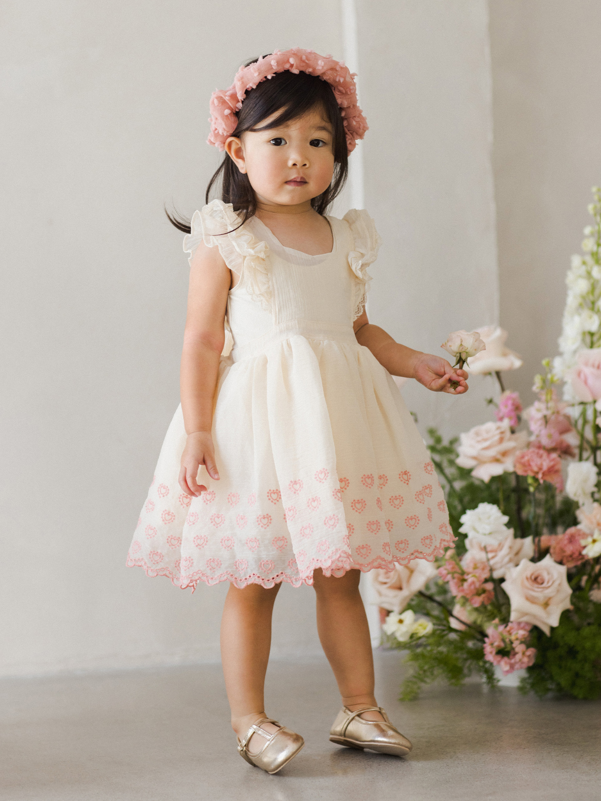 toddler in a cream dress with pink accents, holding a flower, surrounded by floral arrangements.
