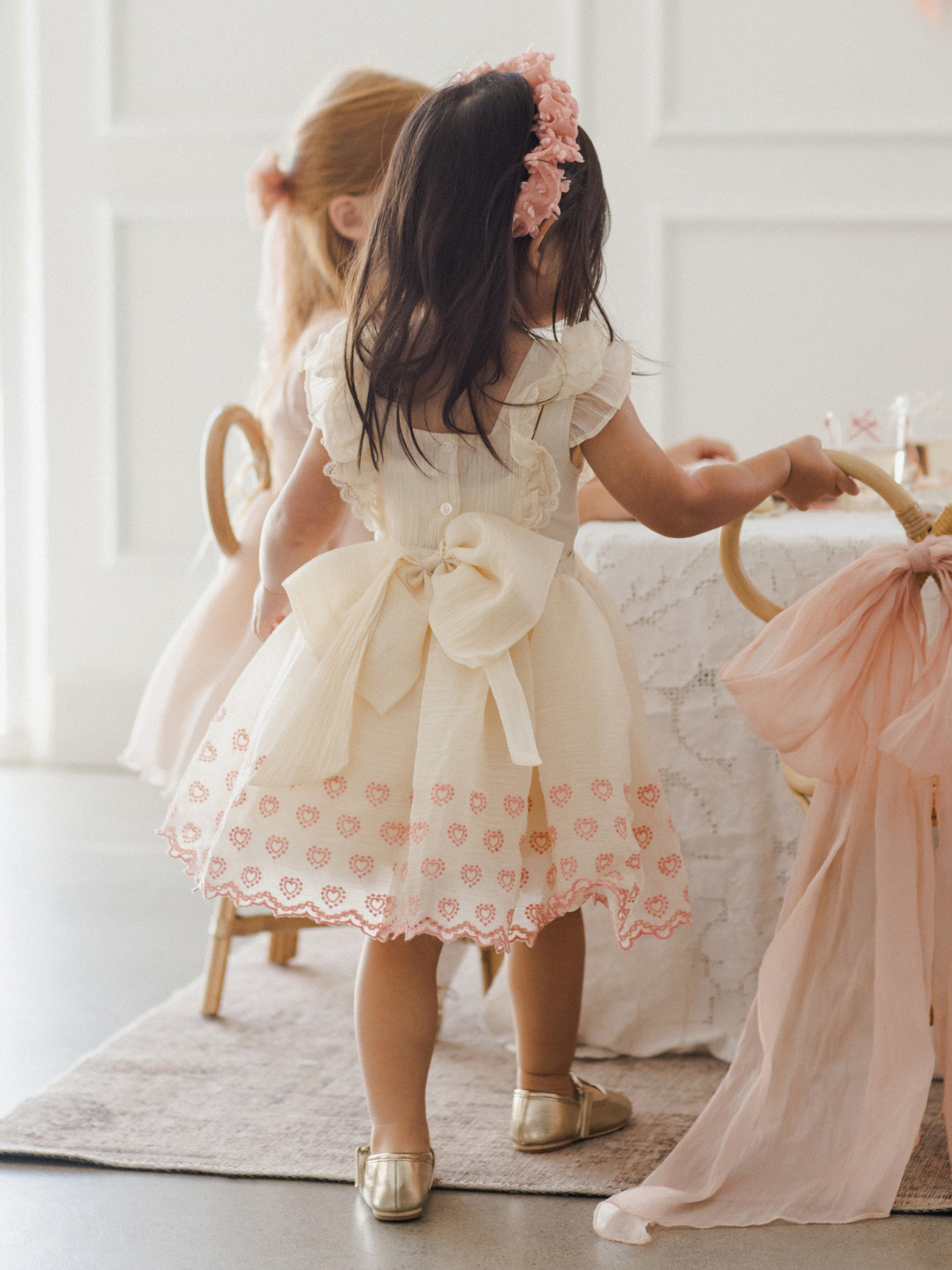 Young girl in a vintage dress with a bow, playing at a table with friends in a bright, stylish setting.