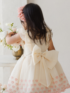 Child in a floral dress with a large bow, arranging flowers on a table. Soft, bright background.