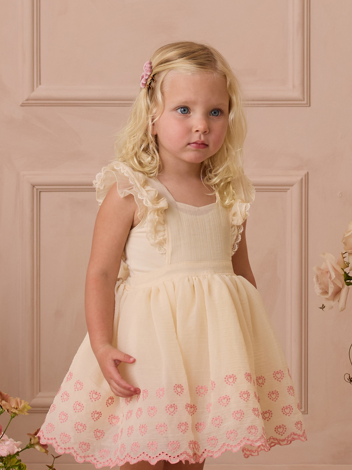 Young girl with blonde, curly hair in a light dress with pink embroidery, posing against a soft pink backdrop.