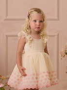 Young girl with blonde, curly hair in a light dress with pink embroidery, posing against a soft pink backdrop.