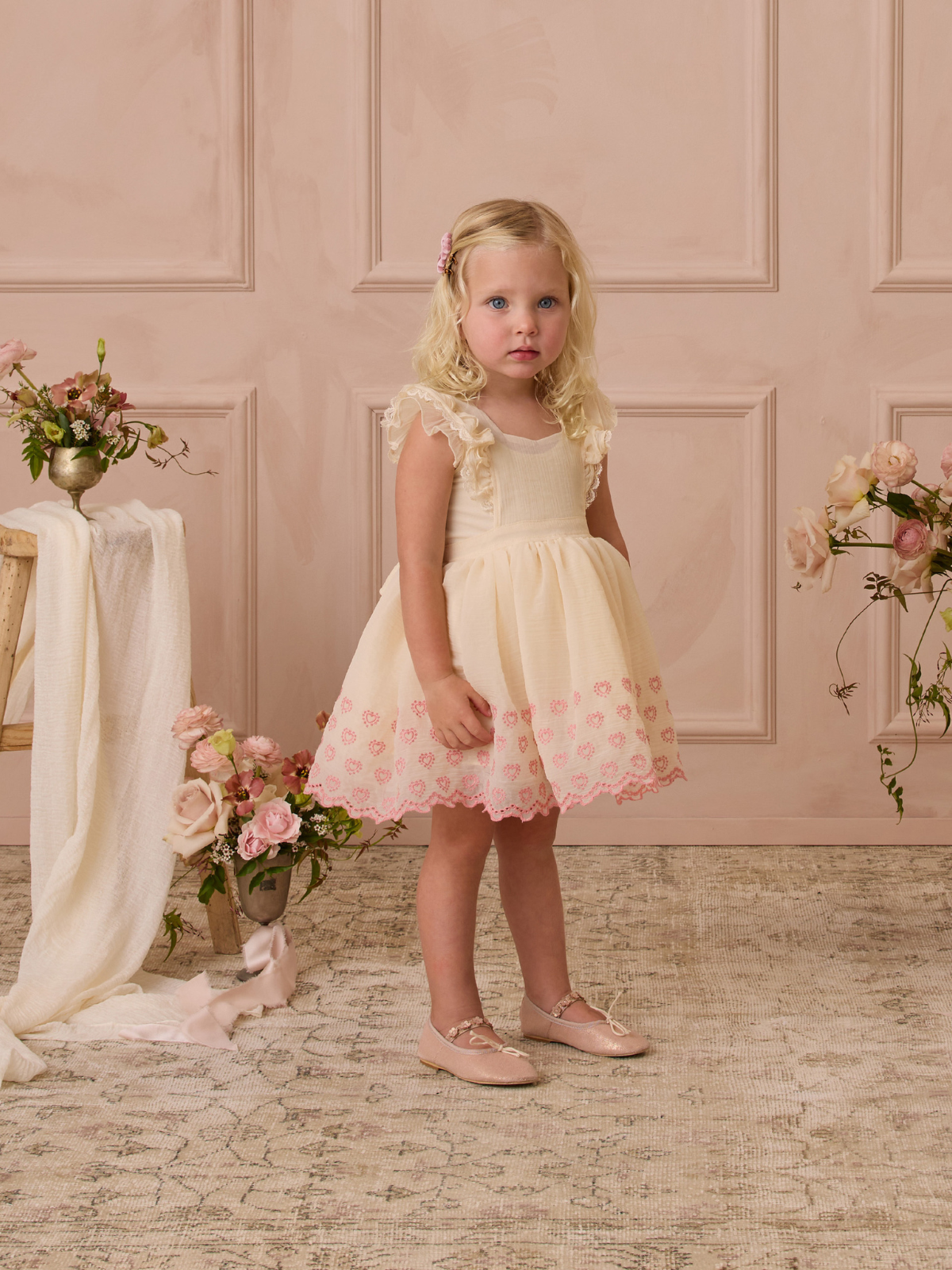 Young girl in a light dress with pink accents, standing in a softly styled room with floral decor.
