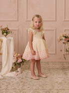 Young girl in a light dress with pink accents, standing in a softly styled room with floral decor.