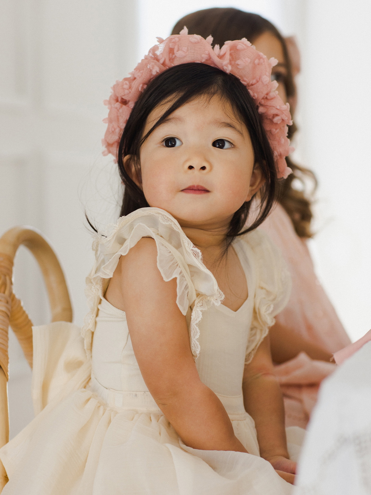 Young girl in a light dress and a pink floral headband, looking curiously at the camera.