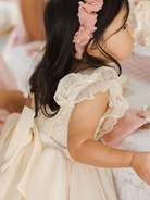 Girl in a cream-colored dress with ruffled sleeves and a pink headband, sitting at a table.