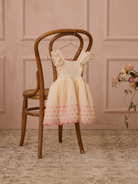 A children's dress with ruffled sleeves hangs on a wooden chair against a soft pink background.