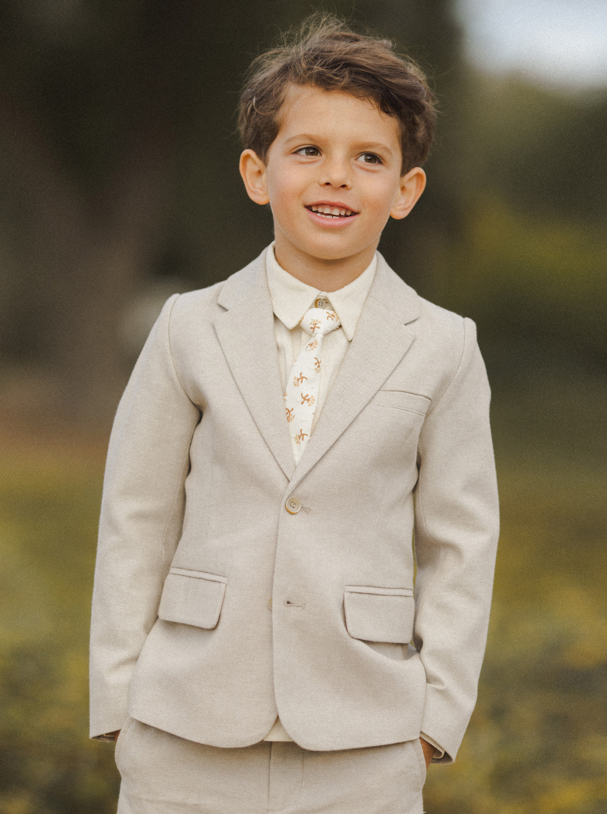 Young boy in a light beige suit with a patterned tie, smiling outdoors.
