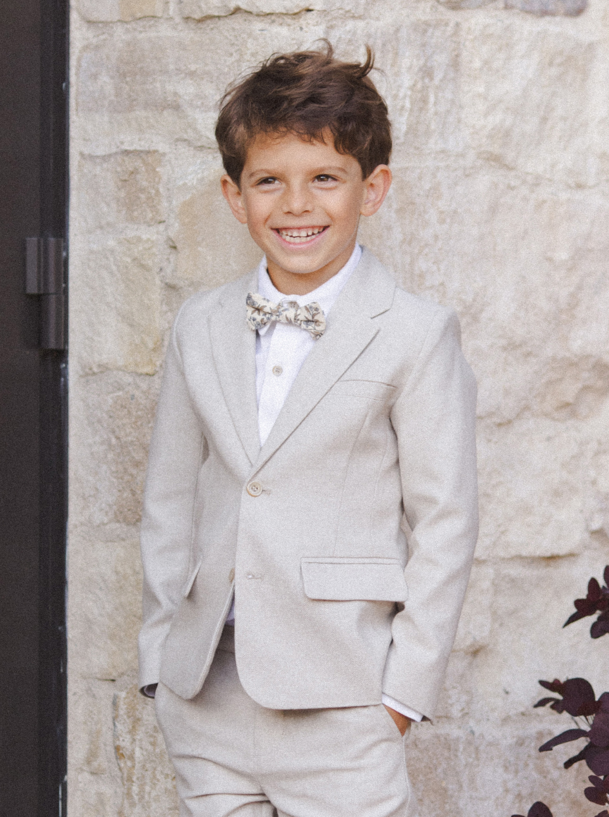 Smiling boy in a light gray suit with a bow tie, standing against a stone wall.