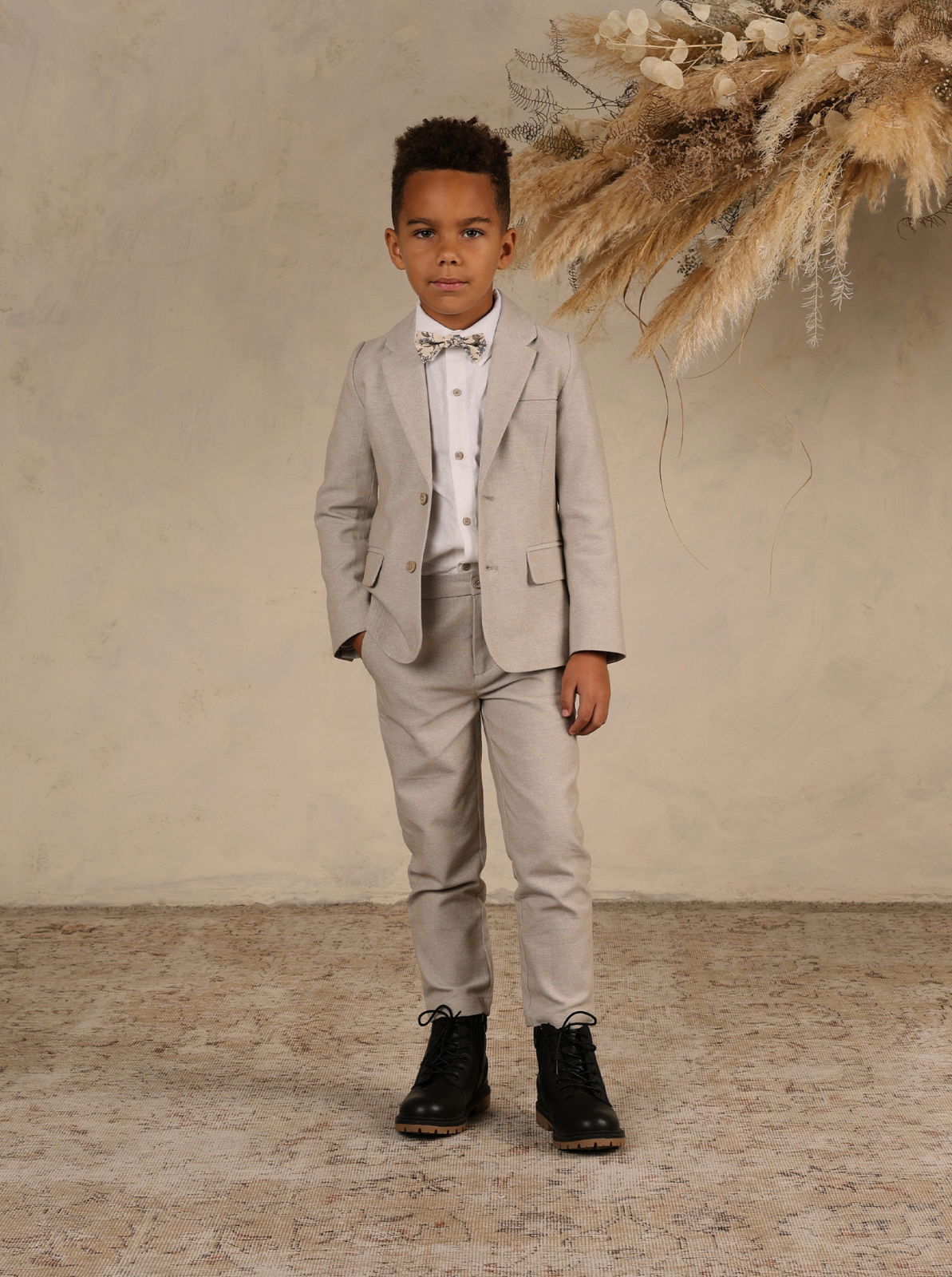Boy in a light gray suit and bow tie, standing confidently against a neutral backdrop with decorative plants.