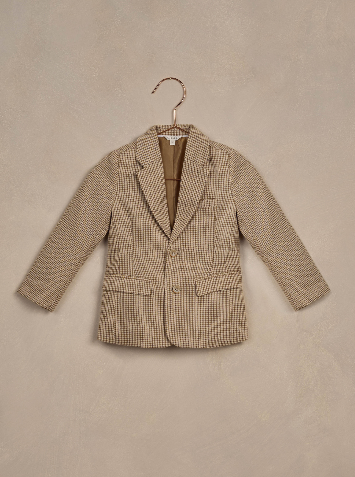 Children's brown plaid blazer hanging on a hook against a textured beige background.