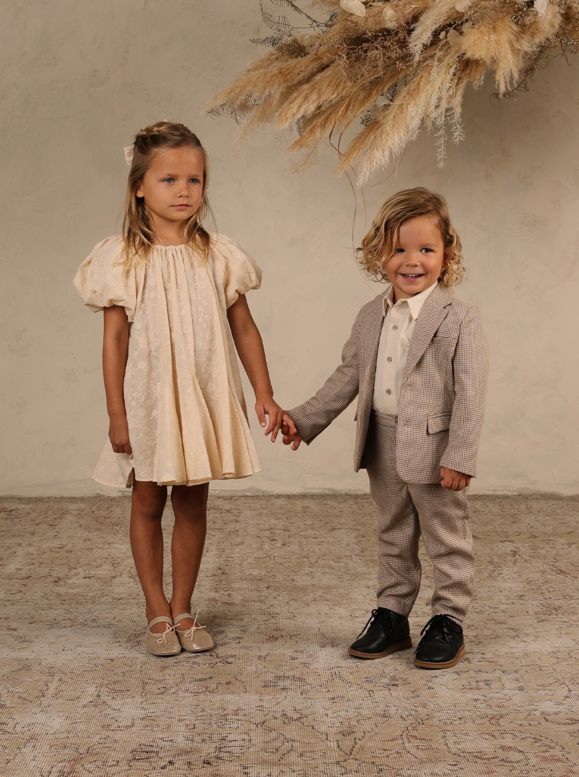 Two children holding hands; a girl in a cream dress and a boy in a patterned suit, smiling against a neutral backdrop.