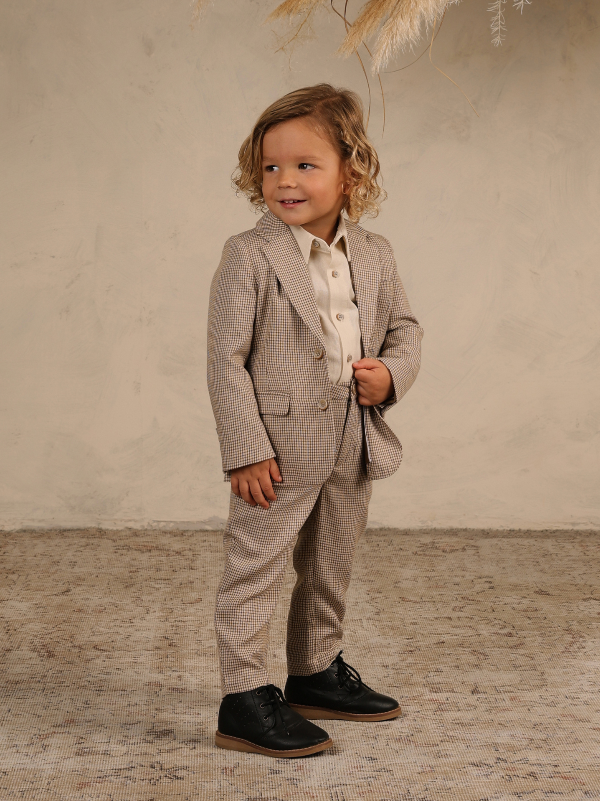 Young boy in a stylish checkered suit with black shoes, smiling against a textured neutral background.