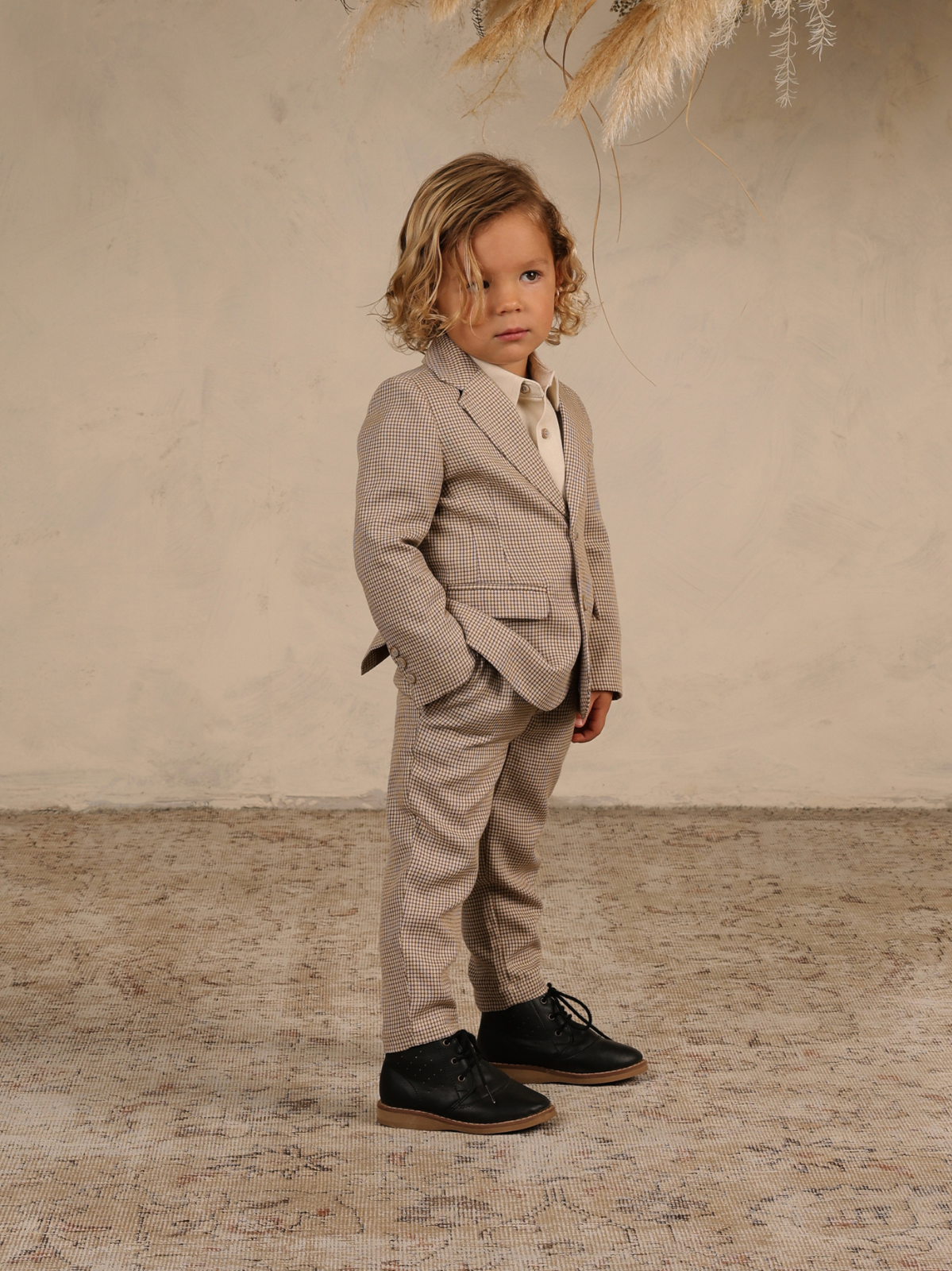 Young boy in a stylish suit stands confidently on a textured rug, with a neutral background and decorative plants.