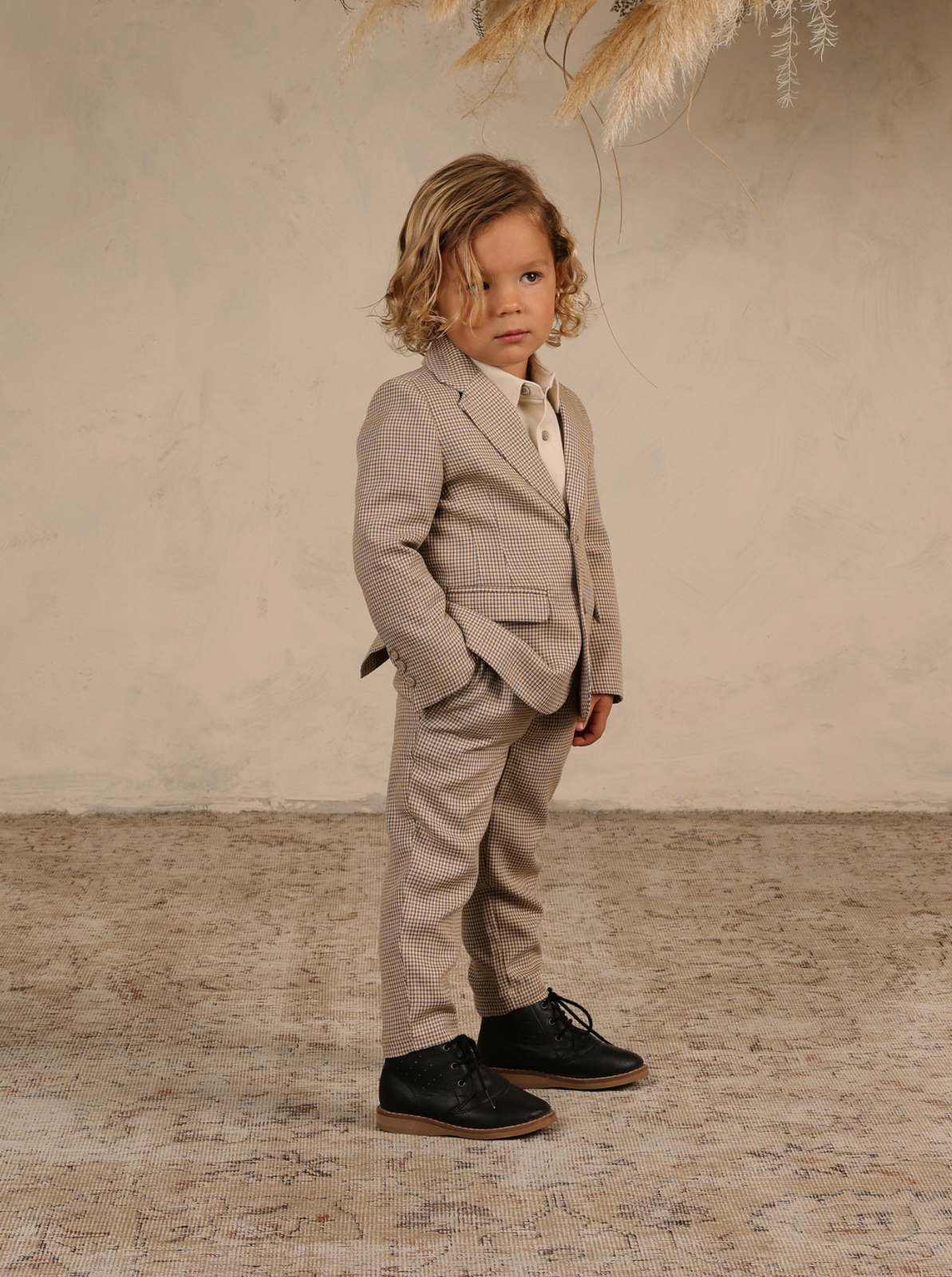 Young boy in a stylish suit stands confidently on a textured rug, with a neutral background and decorative plants.