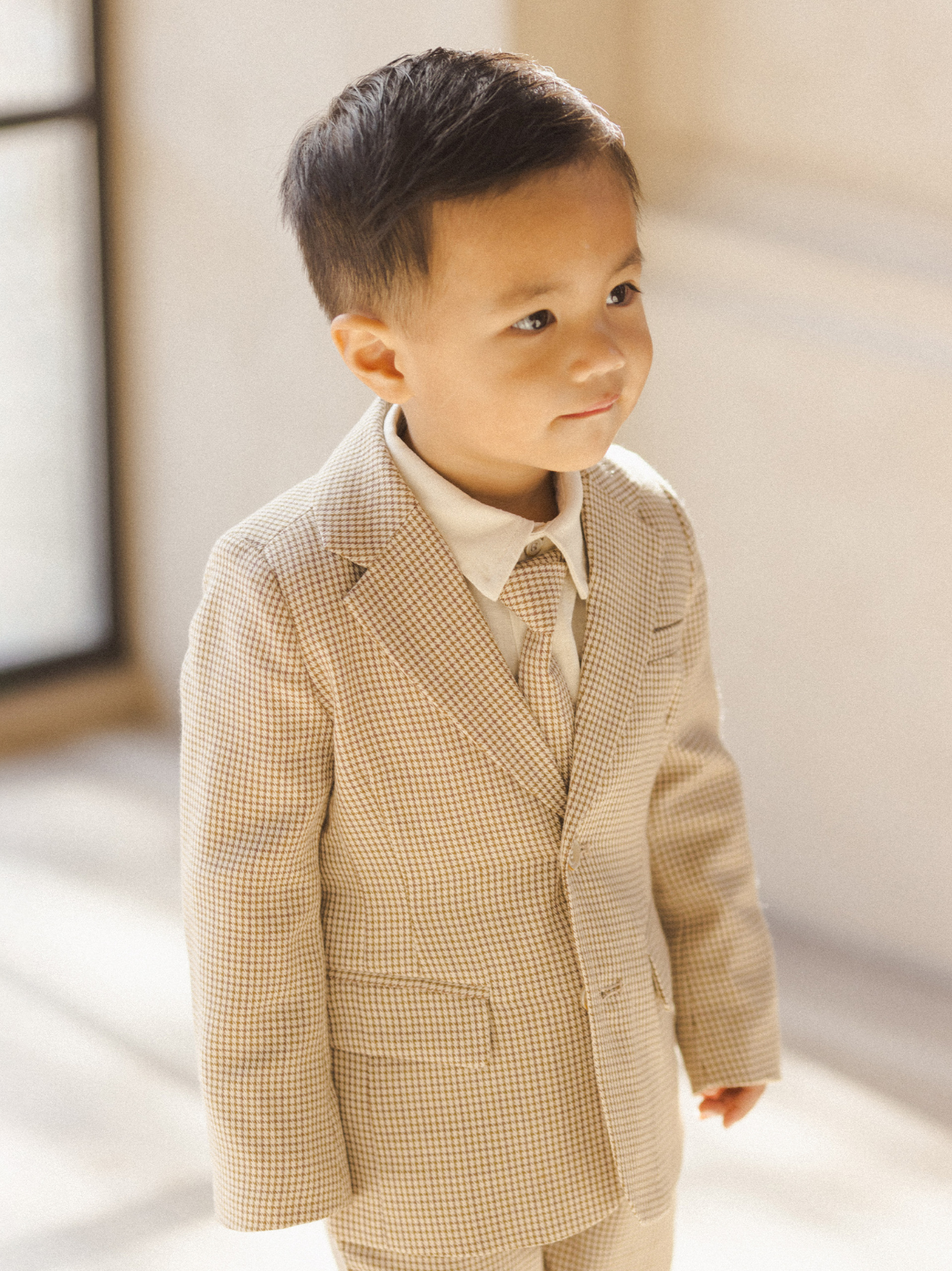 A young boy in a beige checkered suit smiles gently, standing in soft natural light.