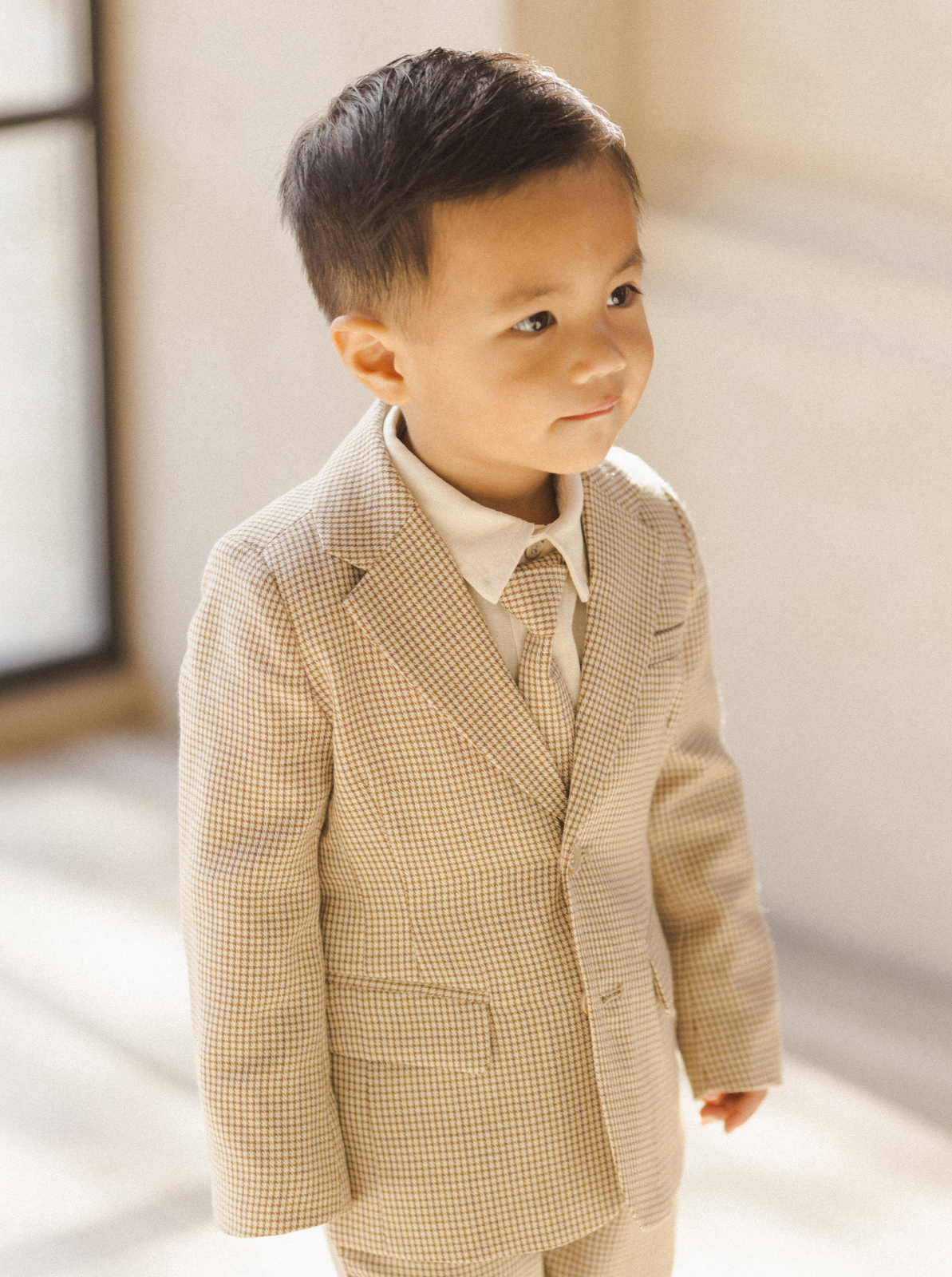 A young boy in a beige checkered suit smiles gently, standing in soft natural light.