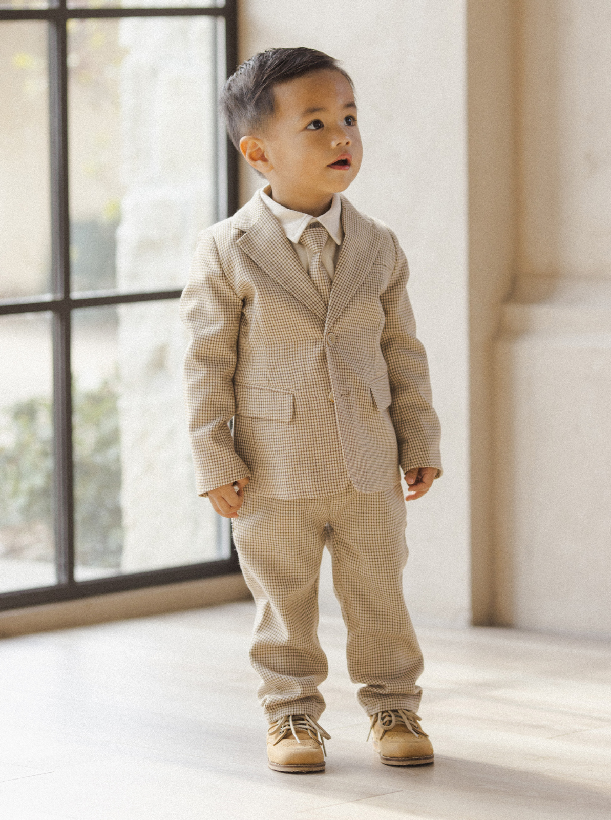 Young boy in a beige checkered suit standing indoors, looking up with a bright expression.