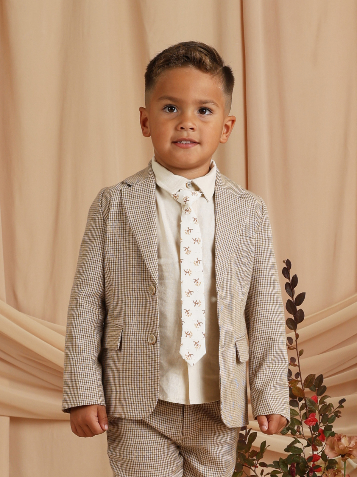 Child in a stylish checked suit with a light tie, smiling against a soft beige backdrop.
