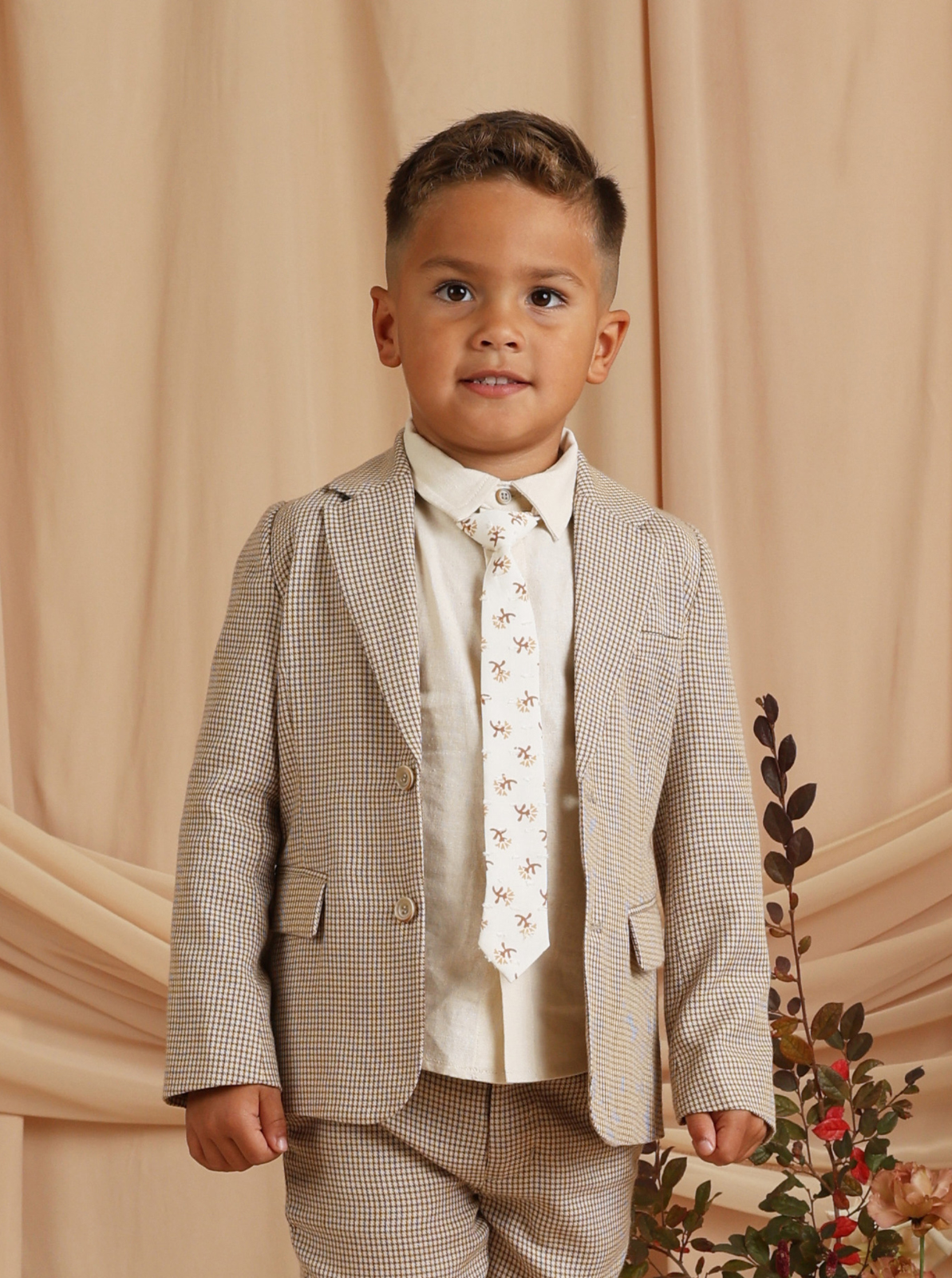 Child in a stylish checked suit with a light tie, smiling against a soft beige backdrop.