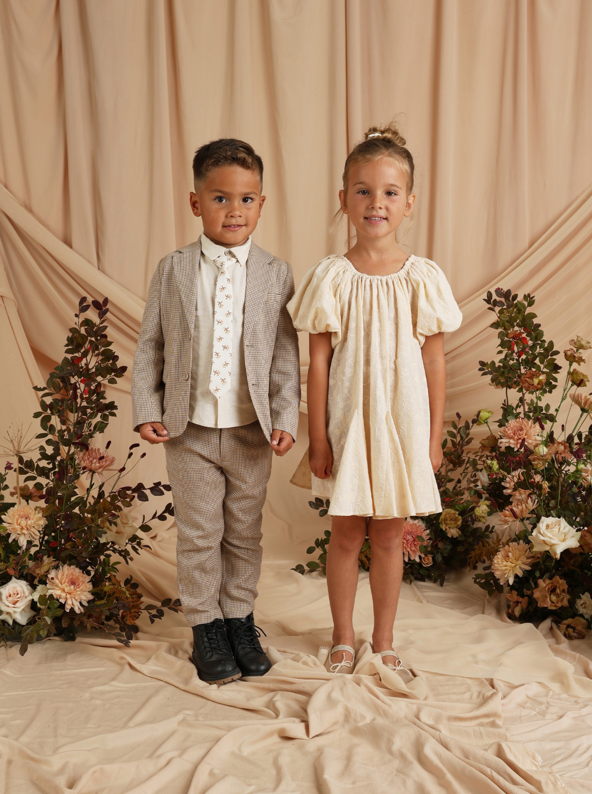 Two children in formal outfits stand posed against a soft beige backdrop with floral arrangements.