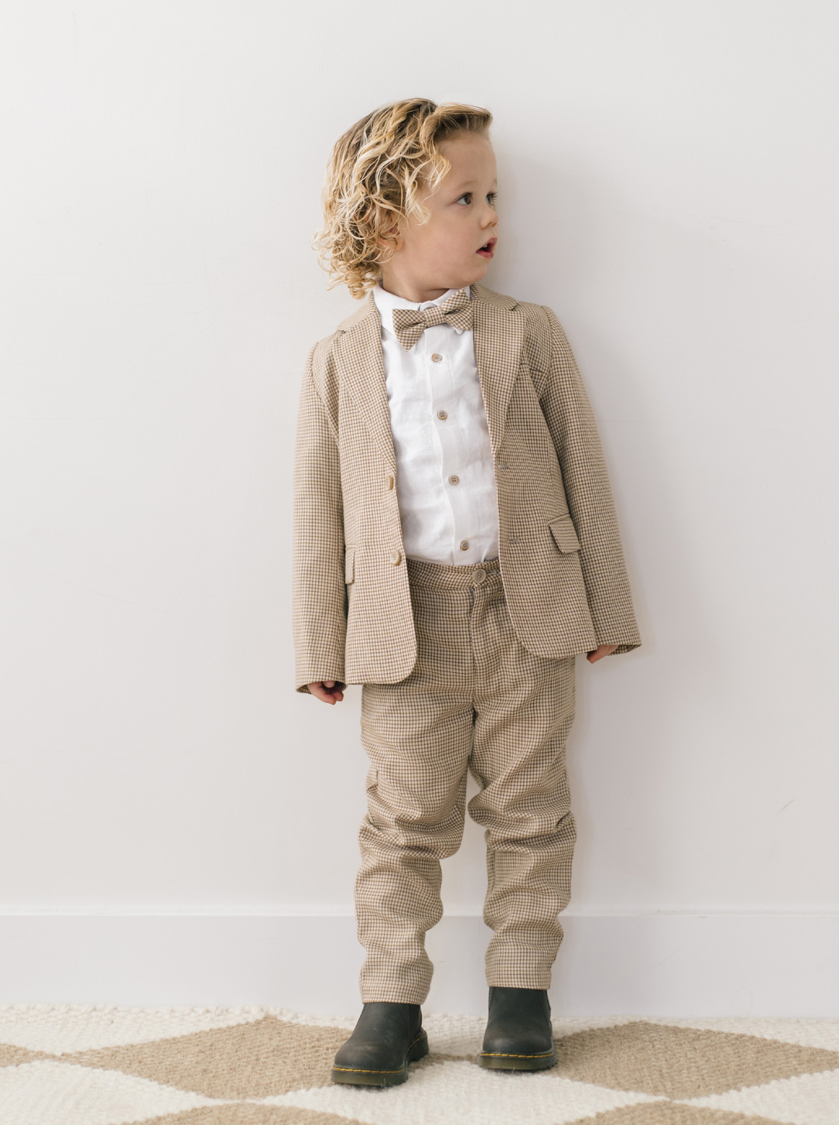 Child in stylish tan suit with bow tie, standing against a white wall, looking to the side.