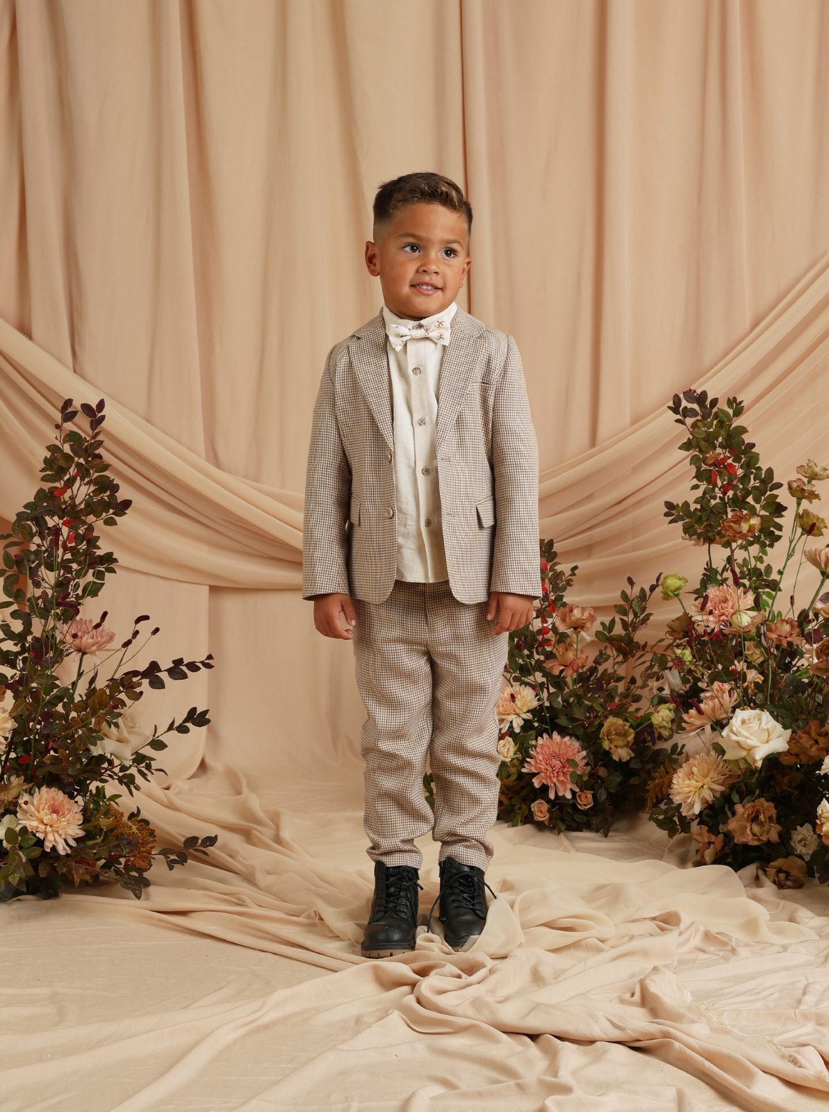 Young boy in a stylish suit with a bow tie, standing amid floral decorations and draped fabric backdrop.