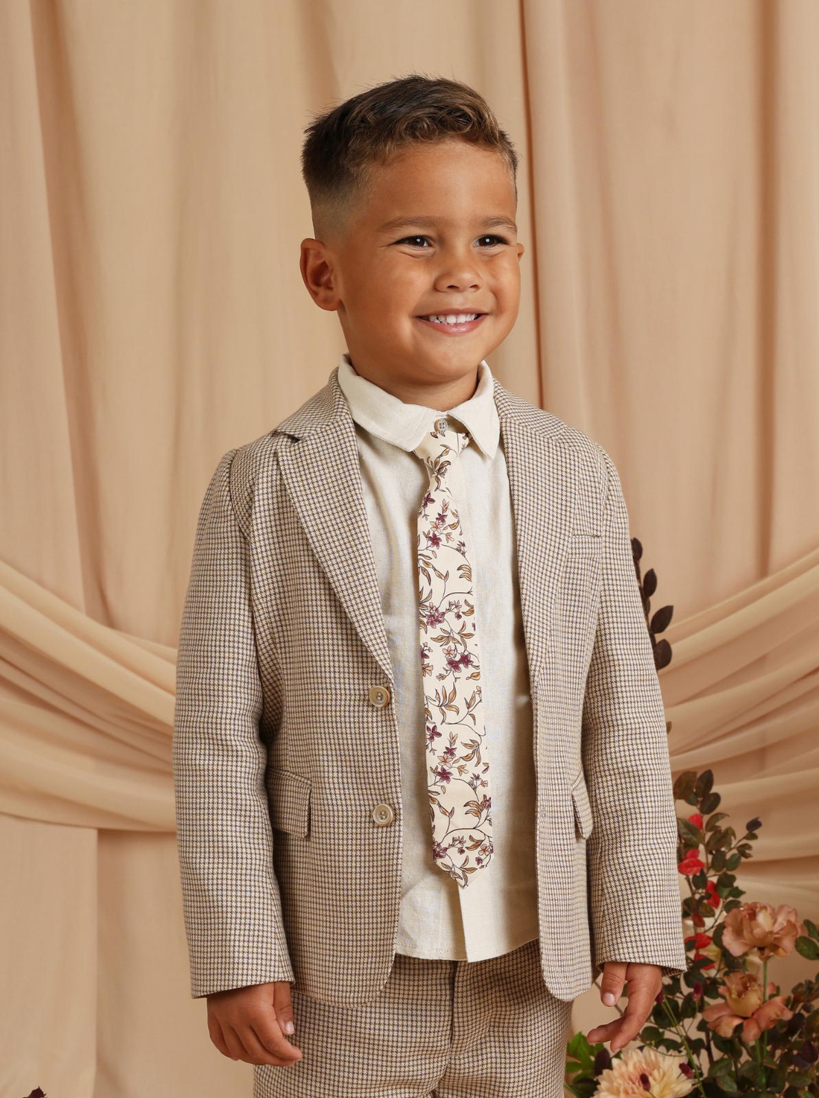 Smiling child in a stylish suit with a floral tie, set against a warm, neutral backdrop.
