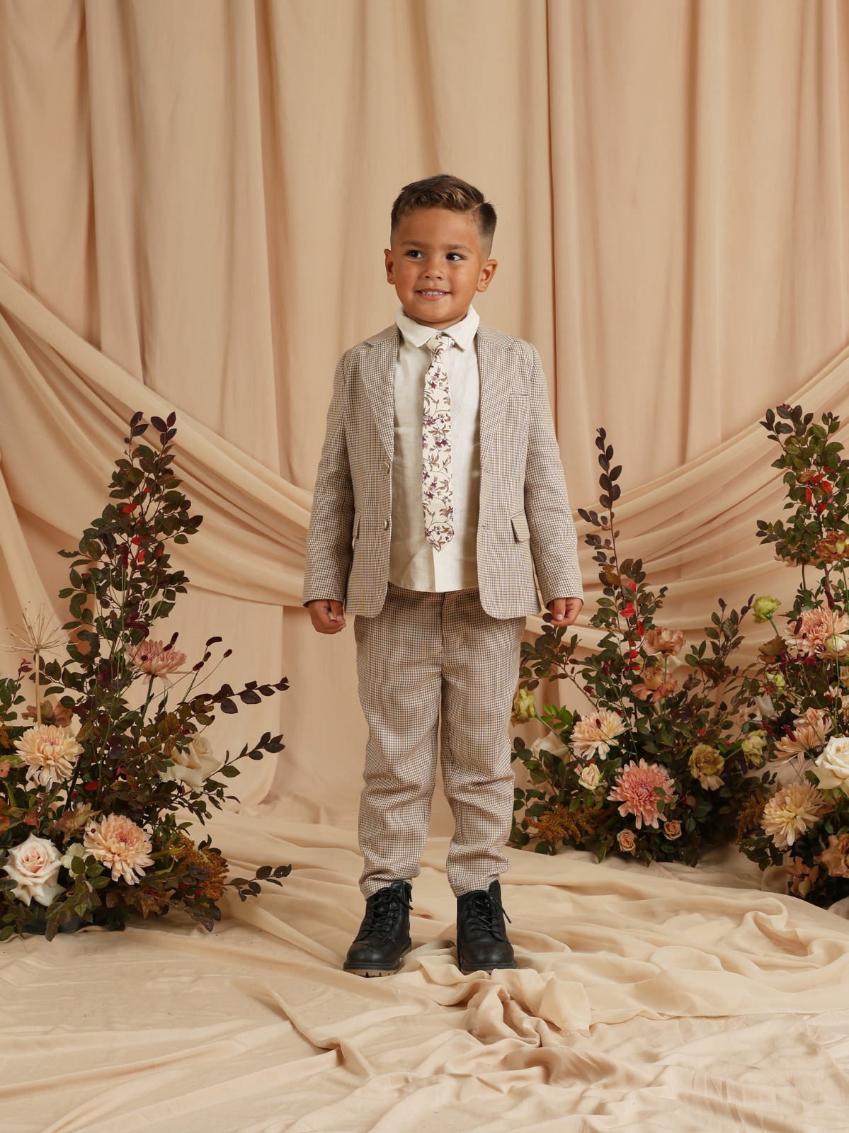 Smiling boy in a stylish suit with floral tie, standing on a neutral backdrop surrounded by flowers.
