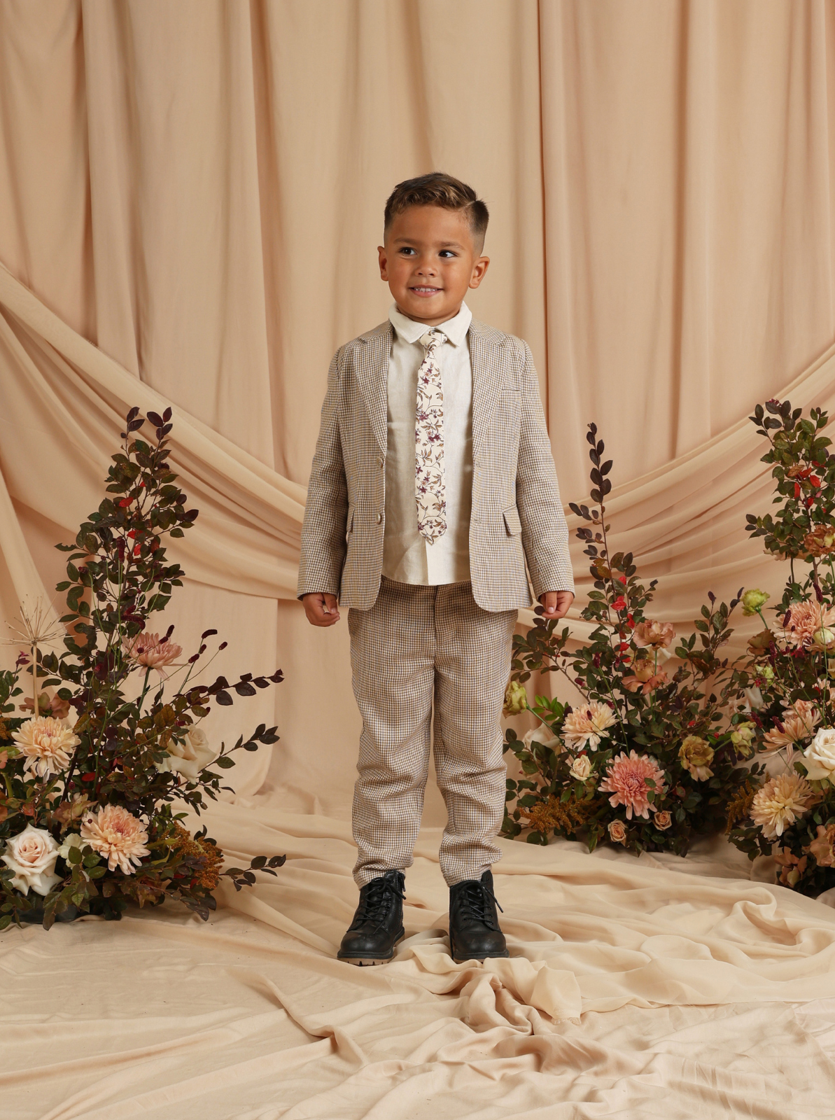 Smiling boy in a stylish suit with floral tie, standing on a neutral backdrop surrounded by flowers.