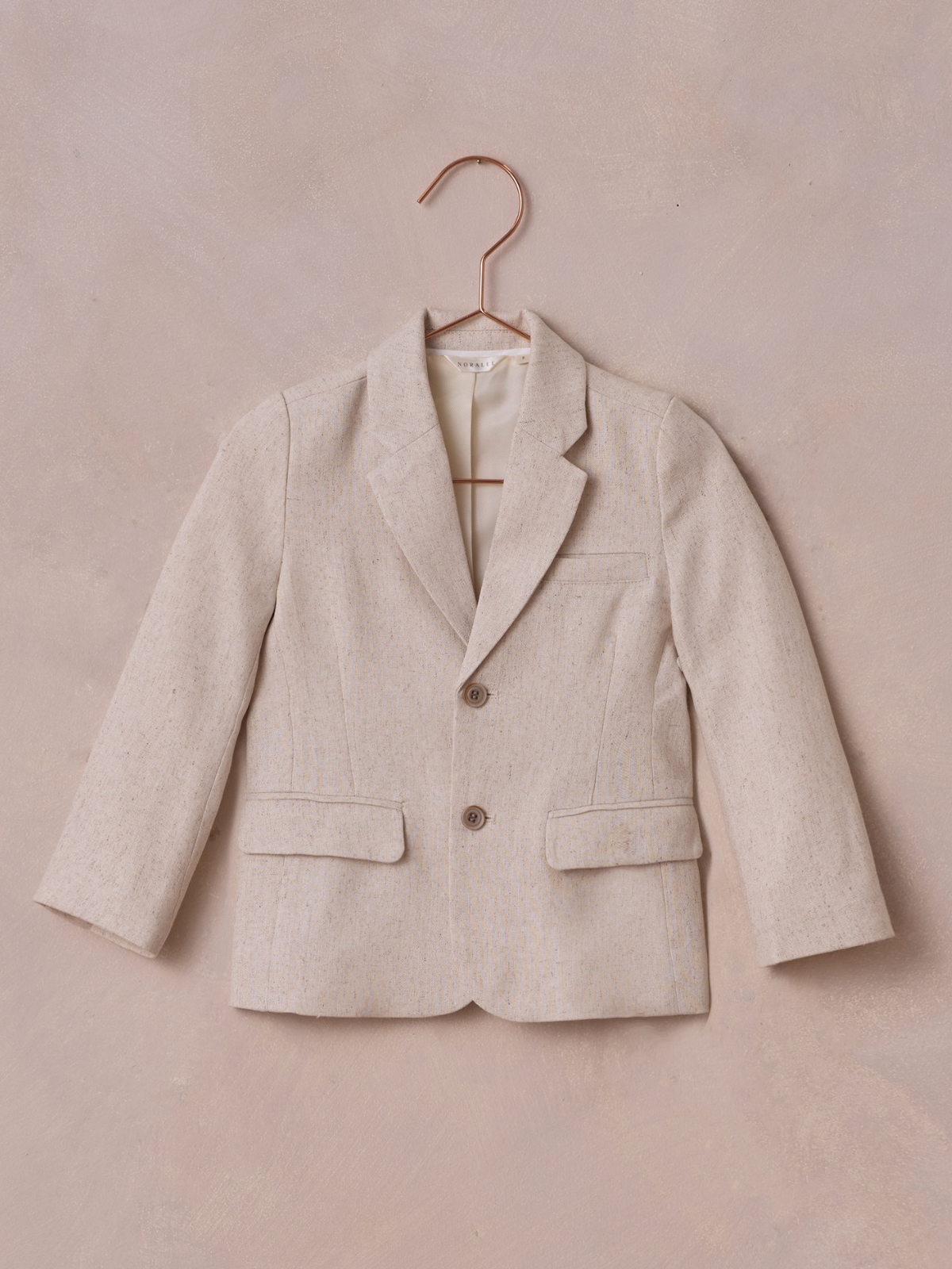 Light beige children's blazer hanging on a rose gold hanger against a textured pink background.