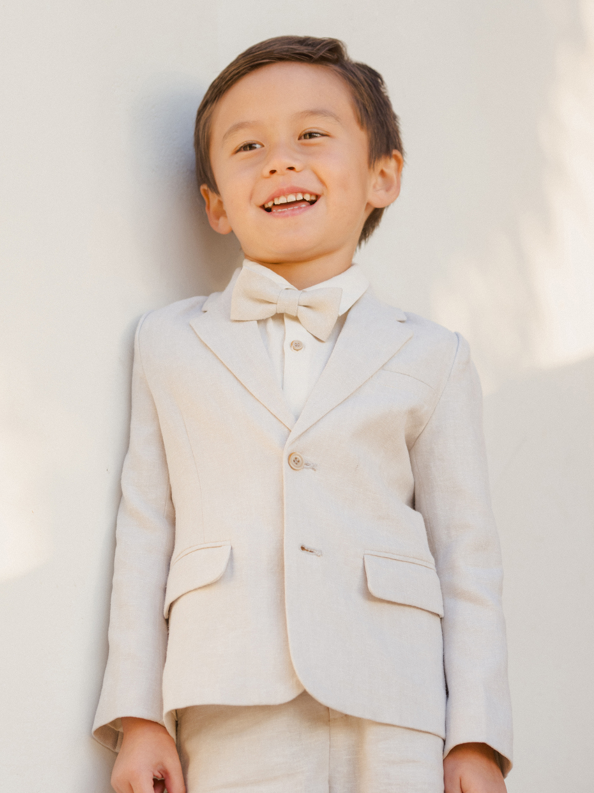Smiling boy in a light-colored suit and bow tie stands against a simple background, radiating joy and style.
