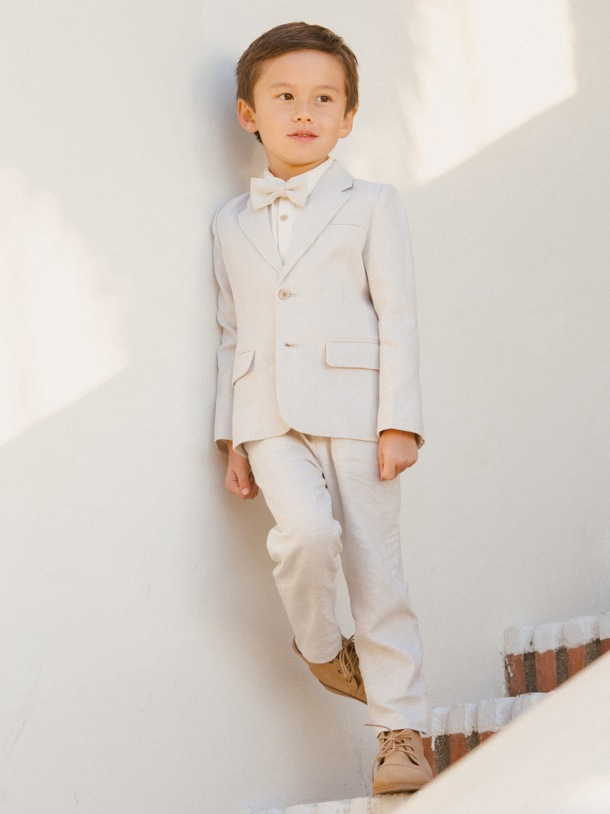 Young boy in a light suit and bow tie, standing on stairs against a pale wall.